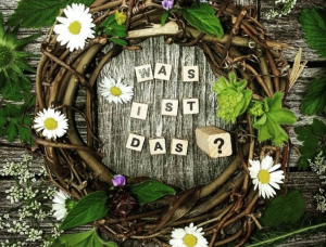 Ein Kranz aus Zweigen, grünen Blättern und weißen Gänseblümchen ist auf einer Holzfläche arrangiert. In der Mitte stehen Scrabble-Kacheln mit der Aufschrift 