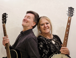 Ein Mann und eine Frau stehen Rücken an Rücken und halten lächelnd jeweils ein Saiteninstrument in der Hand: der Mann eine Gitarre, die Frau ein Banjo mit Sterneinlagen. Sie stehen vor einem schlichten, hellen Hintergrund.