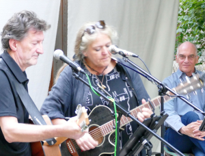 Zwei Personen spielen Akustikgitarren und singen in Mikrofone, während eine dritte Person in der Nähe sitzt und lächelnd zusieht. Sie befinden sich im Freien vor einem weißen Hintergrund mit Grünpflanzen im Hintergrund.