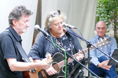 Zwei Personen spielen Akustikgitarren und singen in Mikrofone, während eine dritte Person in der Nähe sitzt und lächelnd zusieht. Sie befinden sich im Freien vor einem weißen Hintergrund mit Grünpflanzen im Hintergrund.