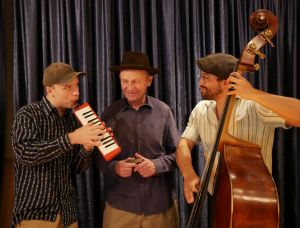 Drei Männer stehen vor dunklen Vorhängen; einer spielt eine Melodica, ein anderer lächelt und hält eine Mundharmonika in der Hand, und der dritte spielt einen Kontrabass. Sie alle tragen legere Hemden und Hüte und genießen einen gemeinsamen musikalischen Moment.
