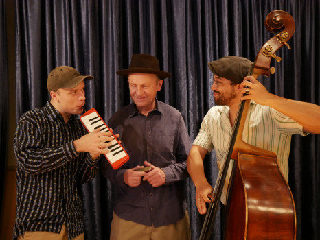 Drei Männer stehen vor dunklen Vorhängen; einer spielt eine Melodica, ein anderer lächelt und hält eine Mundharmonika in der Hand, und der dritte spielt einen Kontrabass. Sie alle tragen legere Hemden und Hüte und genießen einen gemeinsamen musikalischen Moment.