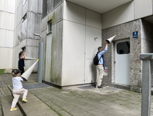 Drei Personen, darunter ein Kind, stehen vor einem modernen Gebäude und halten jeweils große weiße Kegel an die Wände und die Tür, als würden sie lauschen. Die Umgebung ist städtisch mit Betonwänden und einer kleinen Rasenfläche.