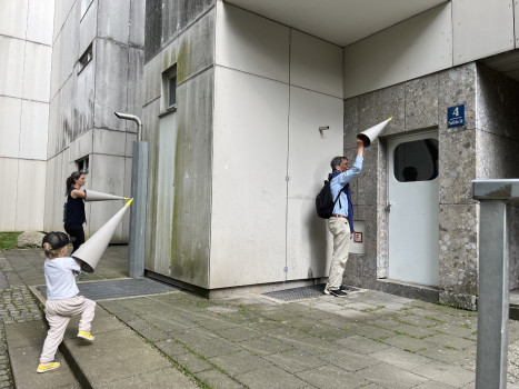 Drei Personen, darunter ein Kind, stehen vor einem modernen Gebäude und halten jeweils große weiße Kegel an die Wände und die Tür, als würden sie lauschen. Die Umgebung ist städtisch mit Betonwänden und einer kleinen Rasenfläche.