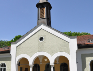 Eine kleine Kirche mit einer hellen Fassade, einem von Säulen getragenen Bogeneingang und einem hohen, mit einem Kreuz gekrönten Kirchturm unter einem klaren blauen Himmel. Auf beiden Seiten des Eingangs sind Topfblumen aufgestellt.