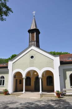 Eine kleine Kirche mit einer hellen Fassade, einem von Säulen getragenen Bogeneingang und einem hohen, mit einem Kreuz gekrönten Kirchturm unter einem klaren blauen Himmel. Auf beiden Seiten des Eingangs sind Topfblumen aufgestellt.