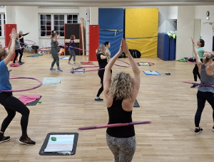 Ein Gruppenfitnesskurs mit Frauen, die in einem Raum trainieren, wobei jede einen Hula-Hoop-Reifen um die Taille trägt. Sie stehen auf einem Holzboden, einige mit erhobenen Armen, und im Raum sind Turnmatten und Handtücher zu sehen.