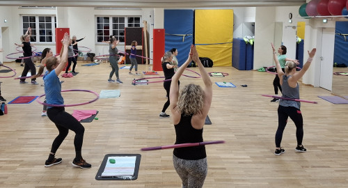 Ein Gruppenfitnesskurs mit Frauen, die in einem Raum trainieren, wobei jede einen Hula-Hoop-Reifen um die Taille trägt. Sie stehen auf einem Holzboden, einige mit erhobenen Armen, und im Raum sind Turnmatten und Handtücher zu sehen.