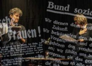 Ein Mann und eine Frau stehen auf der Bühne mit Textblättern in der Hand. Über sie sind Zeitungsausschnitte projiziert. 